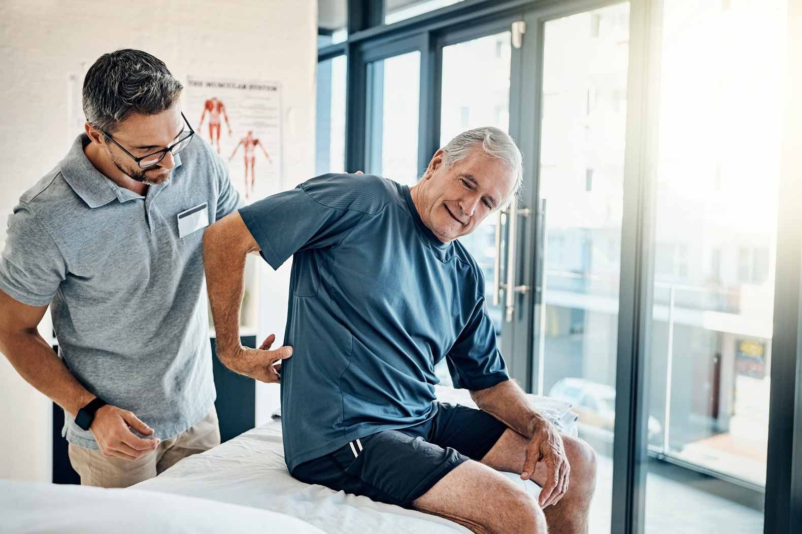 Man helping a seated man adjust his leg during a physical therapy session by a bright window