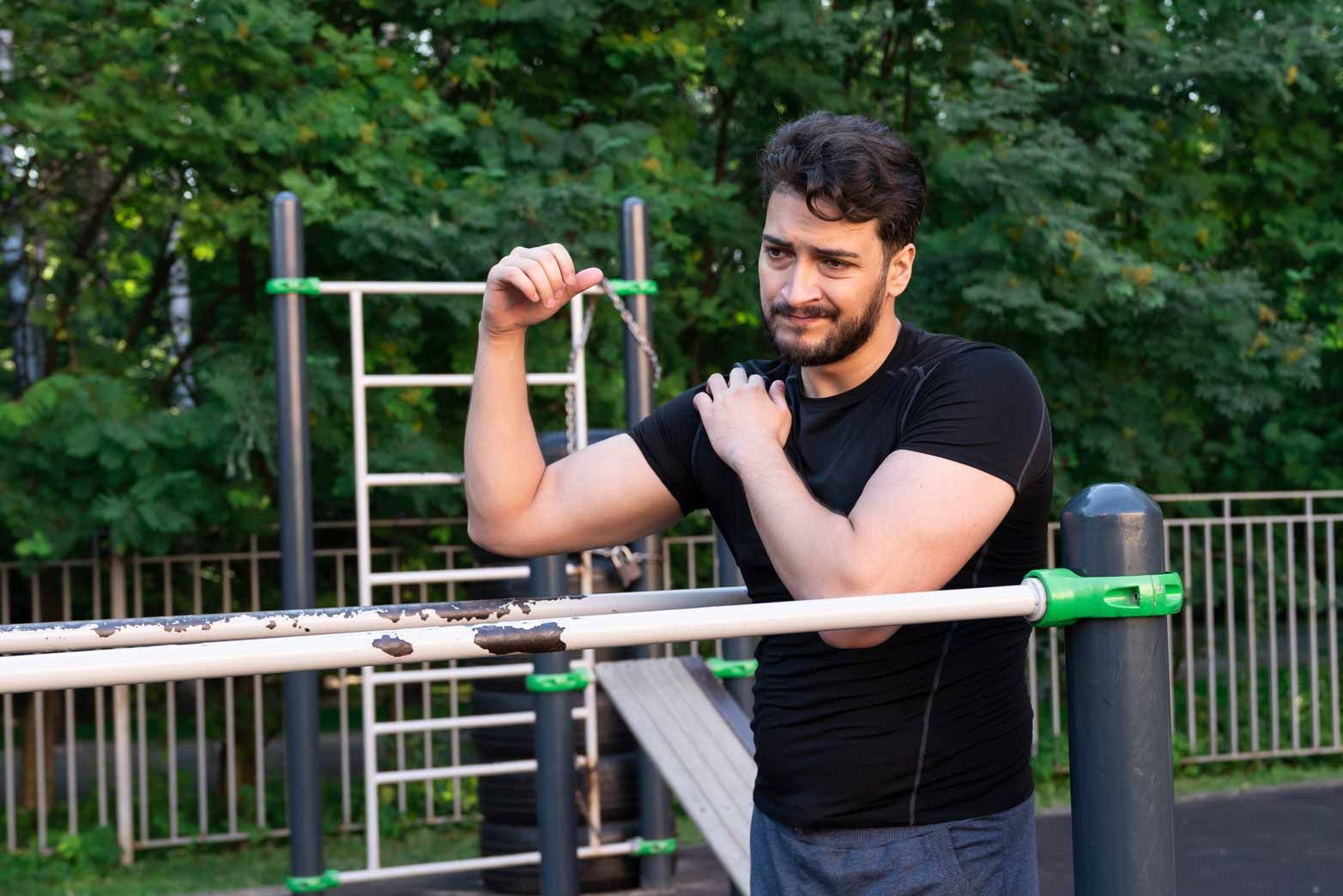 Man in a black shirt flexing by outdoor exercise bars in a park