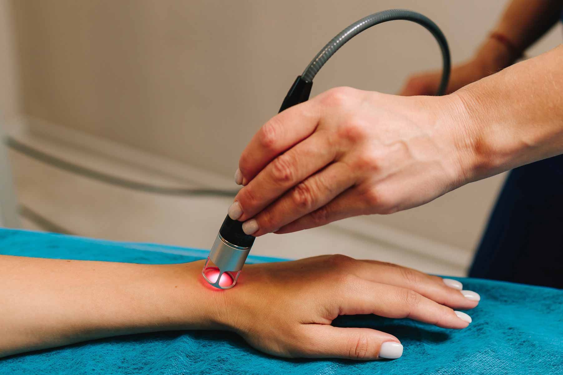 Hand using a handheld laser device on a person’s wrist over a blue towel