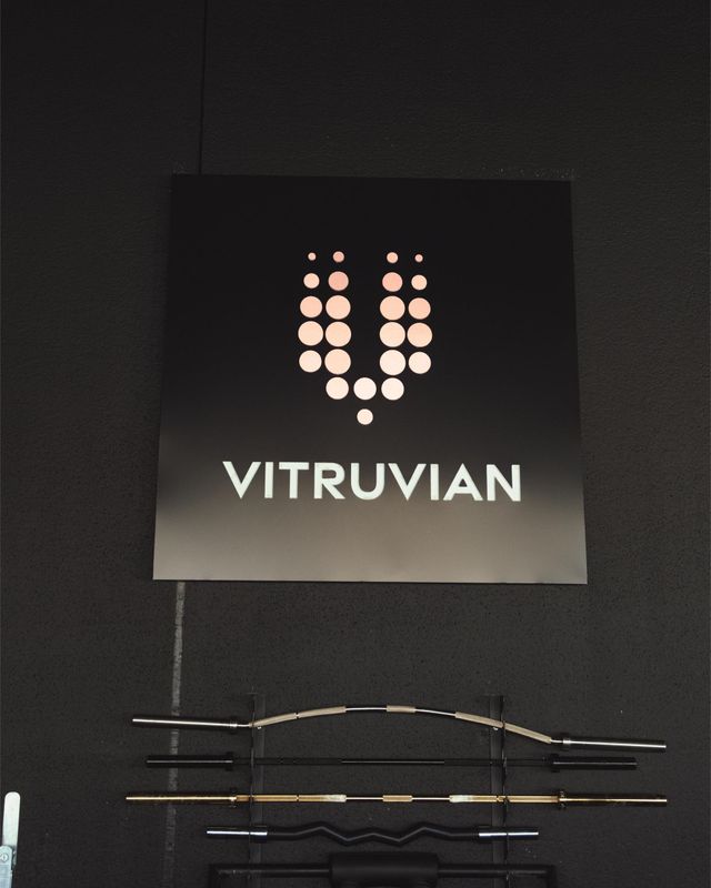Cartello nero con logo a forma di U tratteggiata e la scritta "VITRUVIAN" sopra dei cavi sospesi su una parete scura.