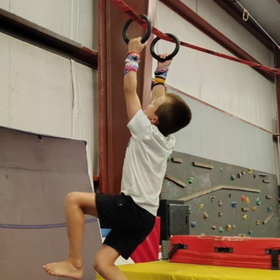 Child hanging from gymnastic rings in an indoor climbing gym, reaching up with both hands.