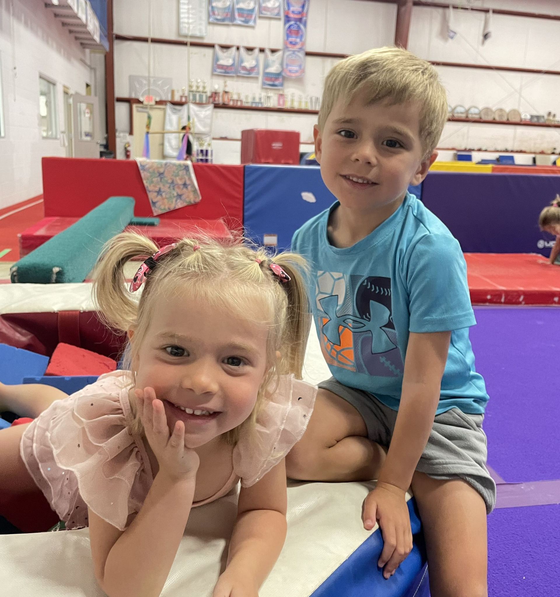 Two children smiling in a gymnastics gym, sitting on colorful mats.