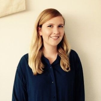 Smiling woman with blonde hair in a navy blouse, standing against a light wall.