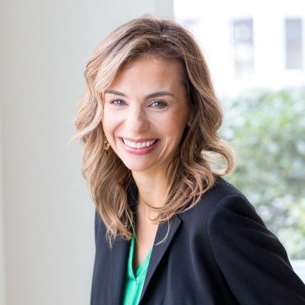 Smiling woman in a black blazer over a green top, standing in a bright office setting
