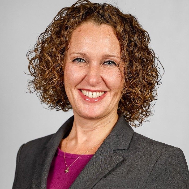 Smiling woman in a gray blazer and purple top against a light gray background