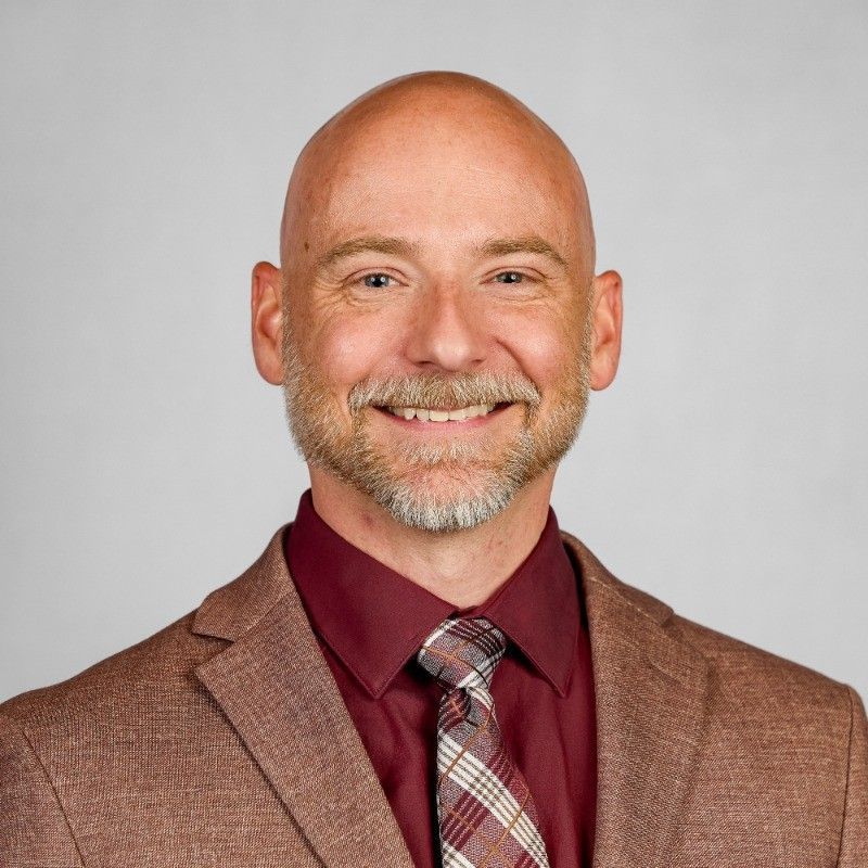 Smiling bald man in a brown suit jacket, maroon shirt, and plaid tie on a gray background
