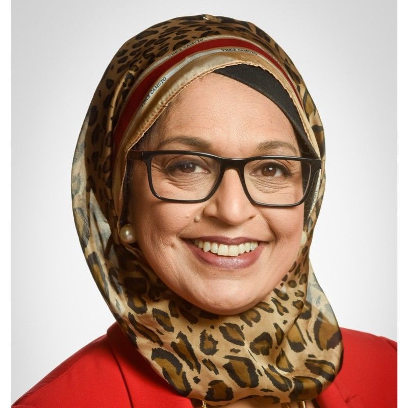 Smiling woman in glasses and patterned hijab wearing a red top against a plain light background