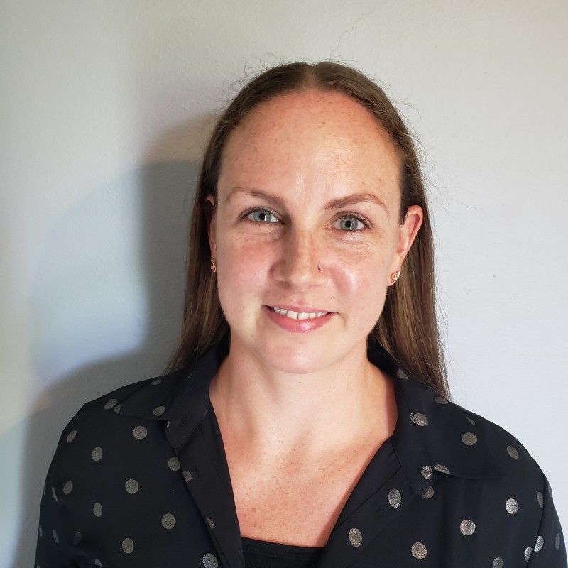 Portrait of a woman with long brown hair, wearing a black polka-dot blouse, against a gray wall