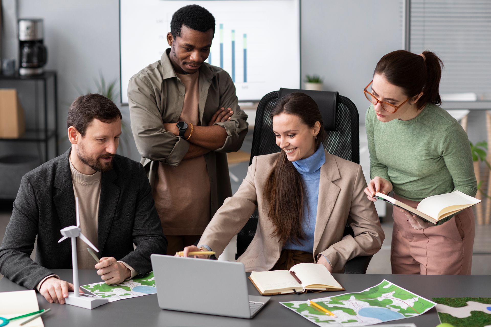A diverse group of professionals collaborates on a sustainability project with a laptop, wind turbine model, and maps.