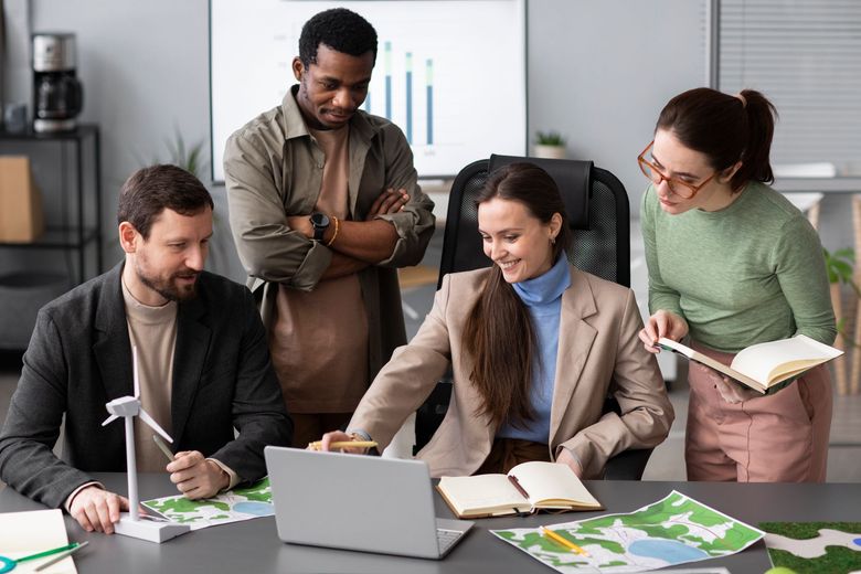 Four coworkers gather around a laptop in a bright office, reviewing charts and documents together.