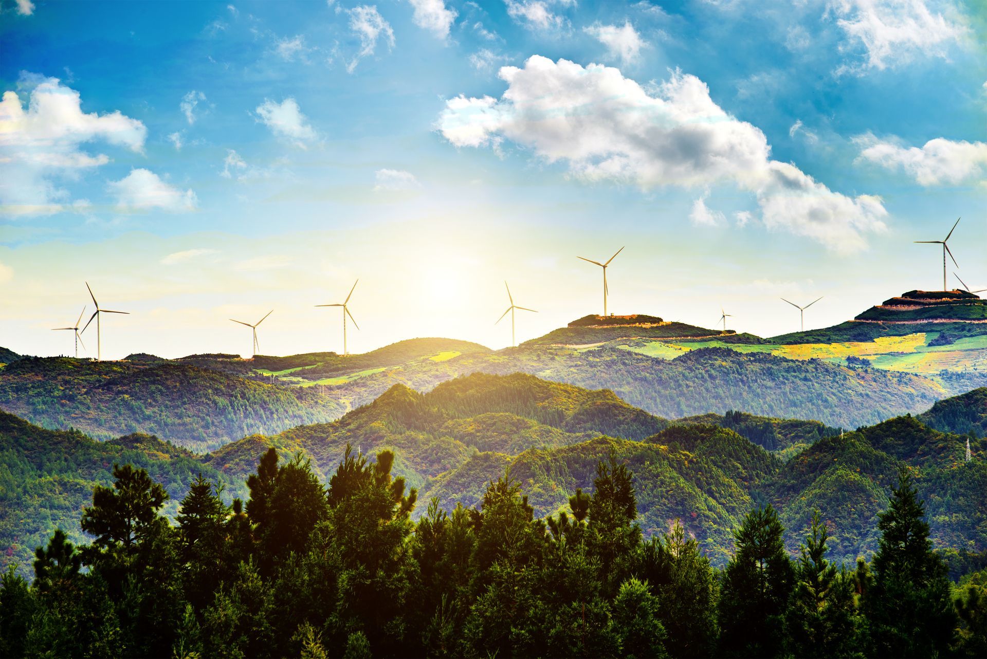 Sunlit mountain ridges with wind turbines and a forest in the foreground