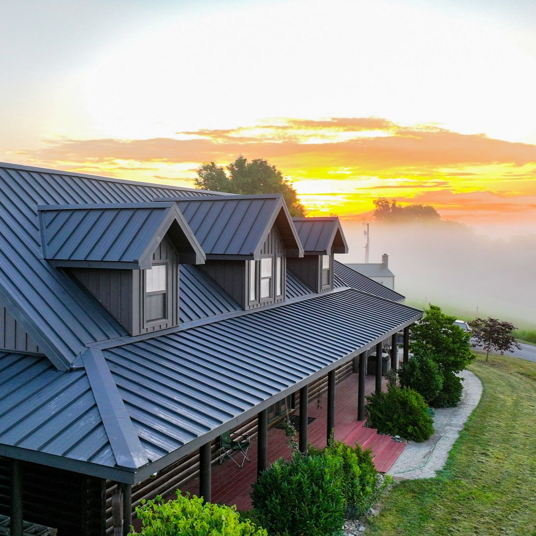 A log cabin with a gray metal roof and three dormer windows sits in a grassy landscape at sunset with fog in the distance.