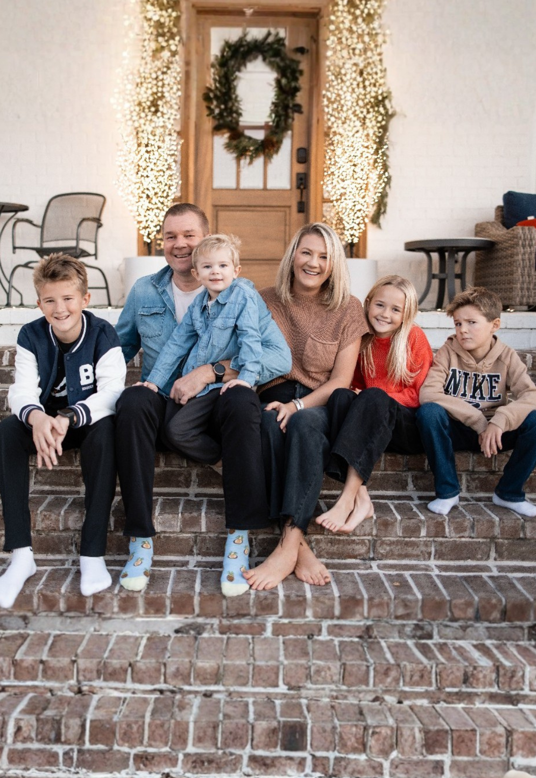 A family of six poses on brick front steps outside a home decorated for the holidays with a wreath and lights.