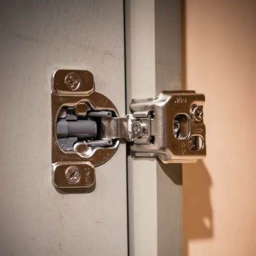 Metal cabinet hinge on a grey cabinet door, attached to a pale orange wall.