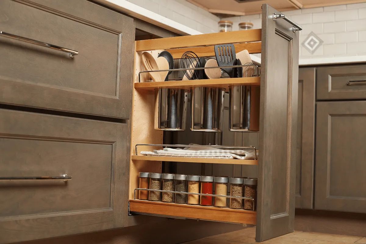Kitchen cabinet with pull-out spice rack, holding spices, utensils, and containers. Gray cabinets.