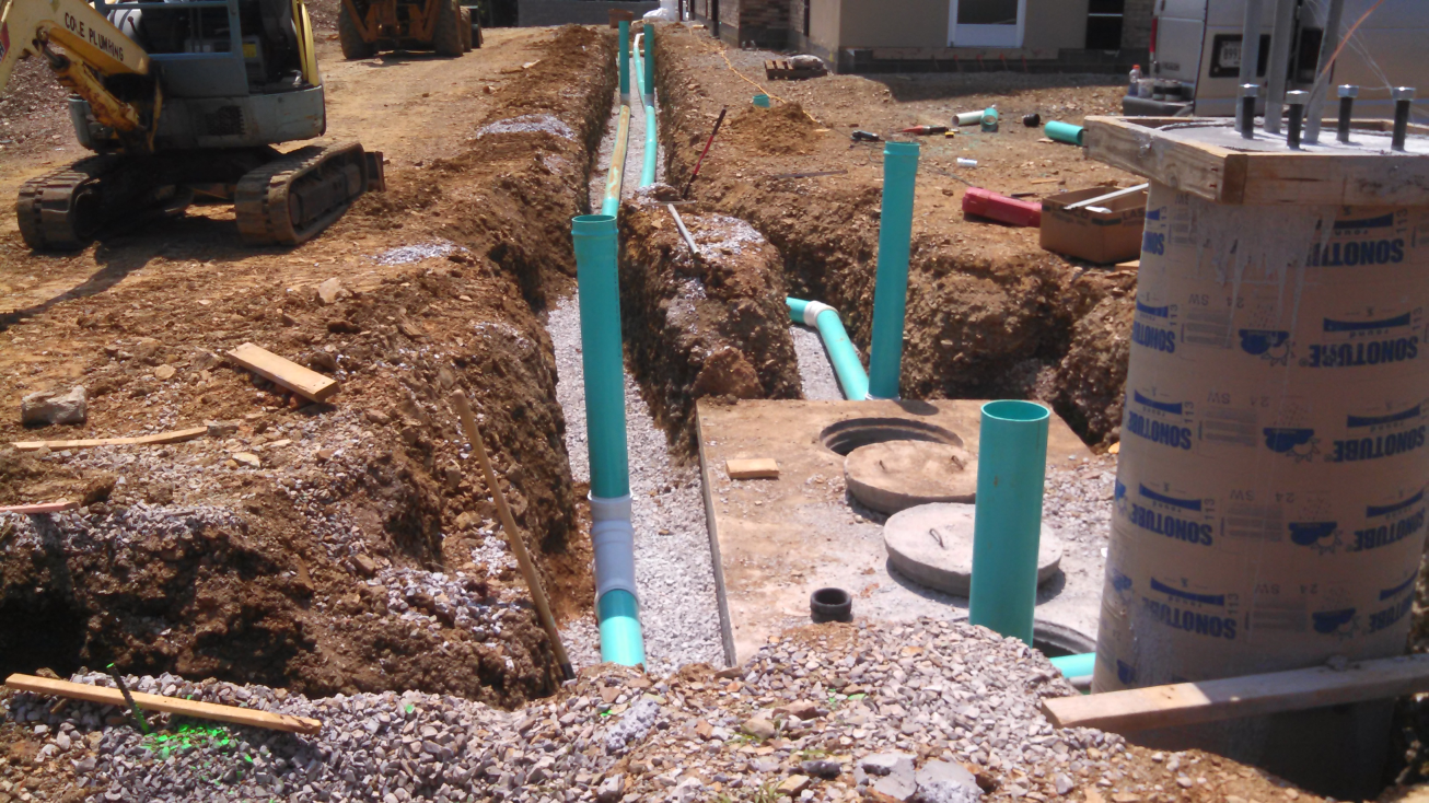Construction trench with exposed pipes and gravel, beside a digger at a residential building site.