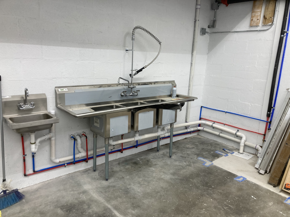 Industrial sink and wash station with stainless counters, white walls, and exposed red and blue plumbing pipes.