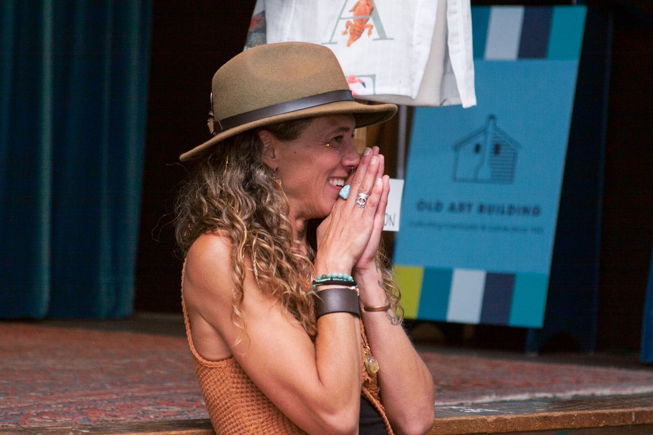 Woman in a tan hat claps with hands near her face on a stage with a blue backdrop.