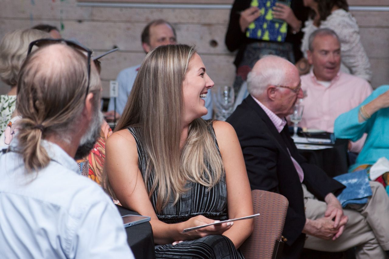 People seated at an event, including a smiling blonde woman in a black dress holding a phone.
