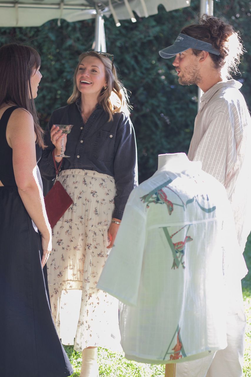 Three people chat outdoors under a canopy, with one holding folded clothing.