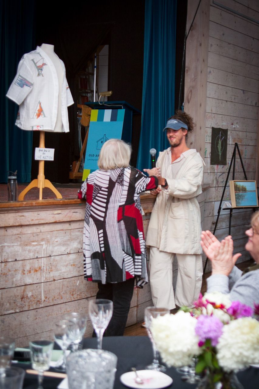Woman hands a red item to another at an indoor stage event, with applauding guests nearby.