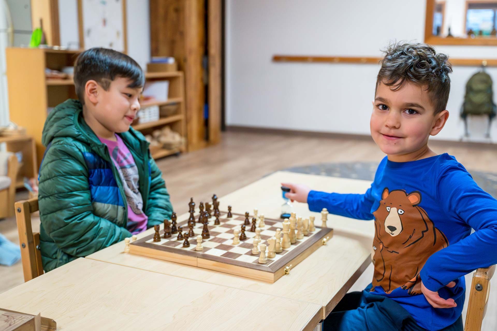 montessori children playing chess at a classroom table with educational shelves in the background