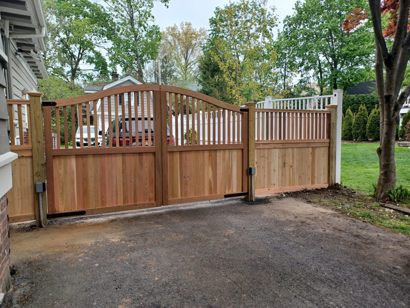 A metal gate stands between stone pillars at the entrance to a gravel driveway leading to a sunny hillside yard.