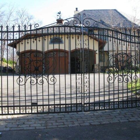 A pair of ornate black wrought iron driveway gates in front of a house with a three-car garage.