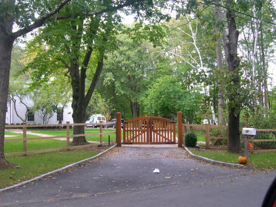 A paved driveway leads to a wooden gate flanked by a split-rail fence, surrounded by green trees and a white house.