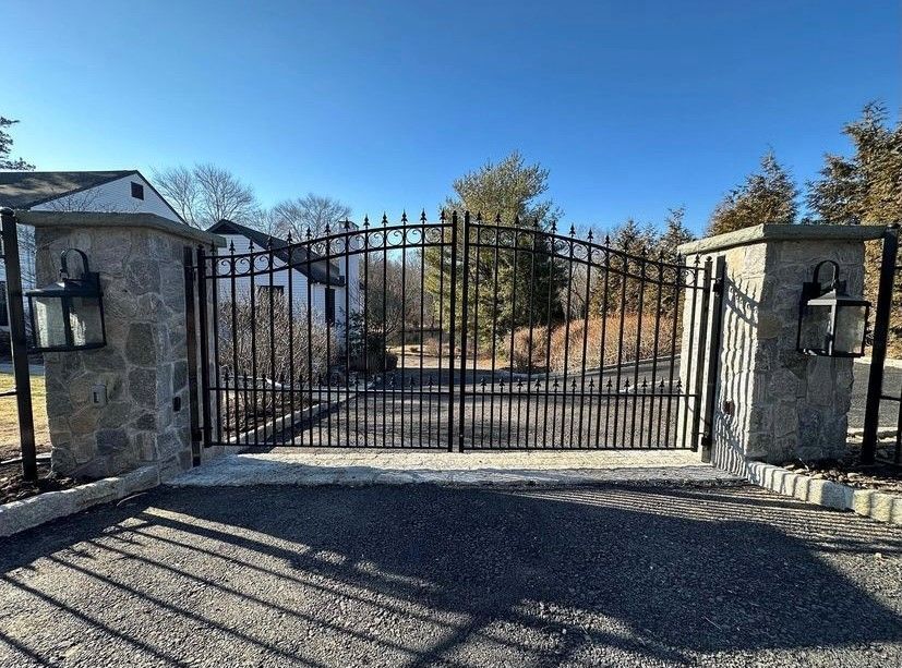 A black metal driveway gate sits between two brick columns, with a light-colored brick house and trees in the background.