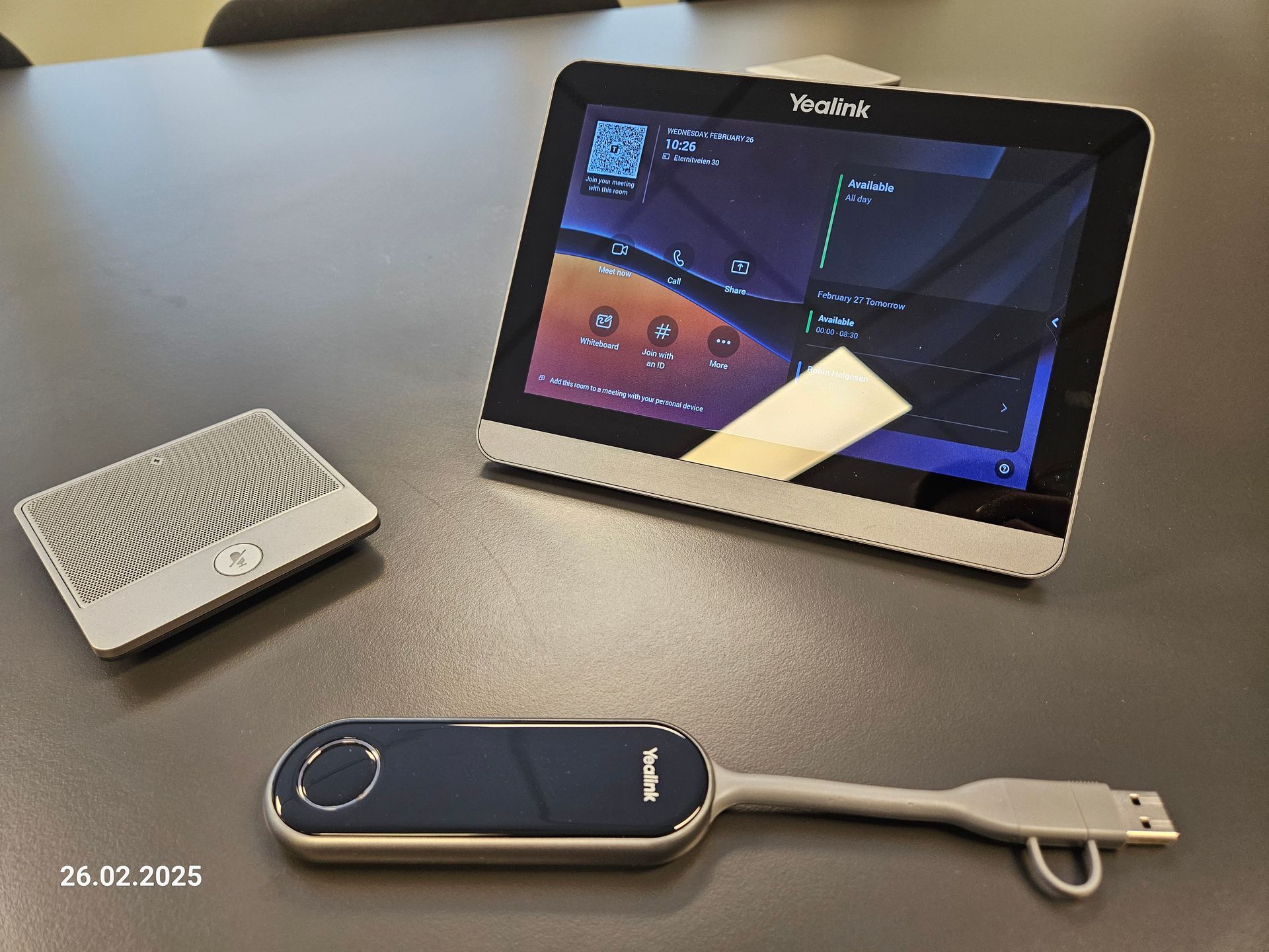 Liten hvit høyttaler, nettbrett på et stativ og en blå USB-stasjon på et grått bord.