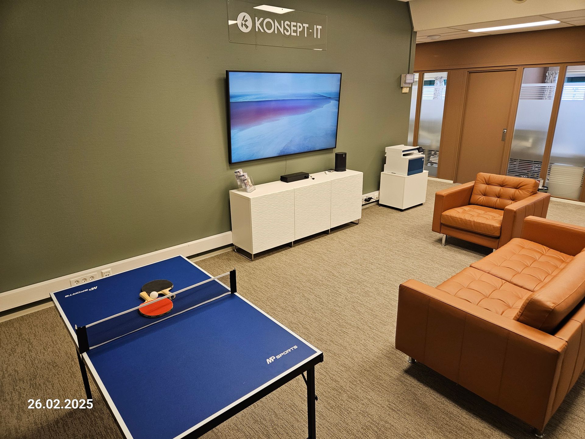 Lys, moderne stue med TV, bordtennisbord og oransje sofaer mot en grå vegg i bakgrunnen.