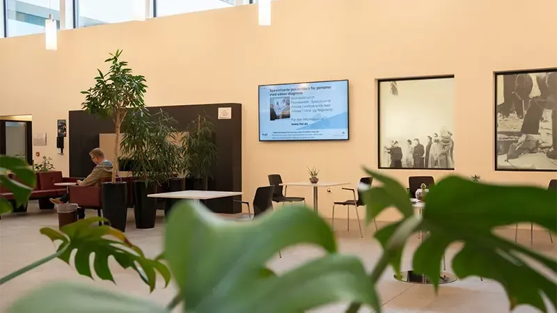 Moderne kontorlobby med sitteplasser, potteplanter, veggkunst og digital informasjonsskjerm