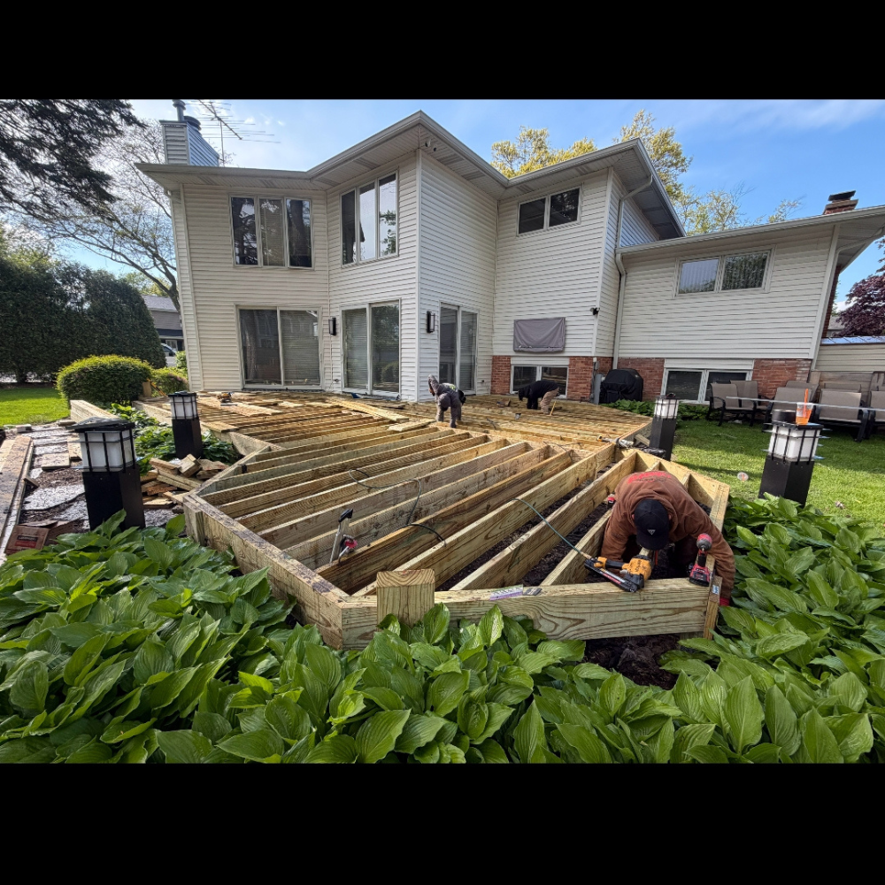 Backyard deck under construction beside a white two-story house, with workers and stacked lumber.