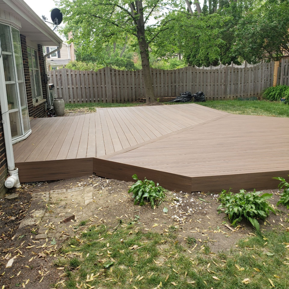 Wooden backyard deck beside a house, bordered by a fenced lawn and trees.