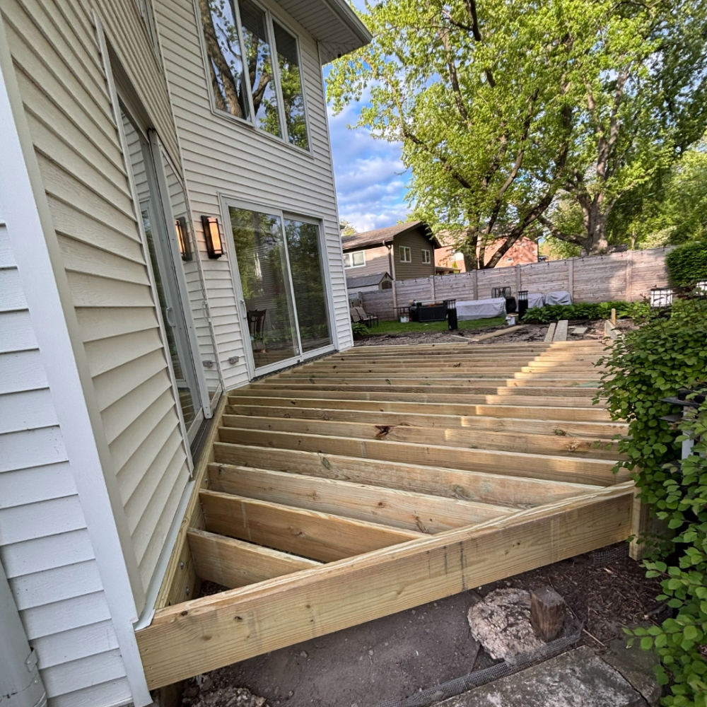 Wooden deck framing under construction beside a house, with joists installed and a tree in the yard.