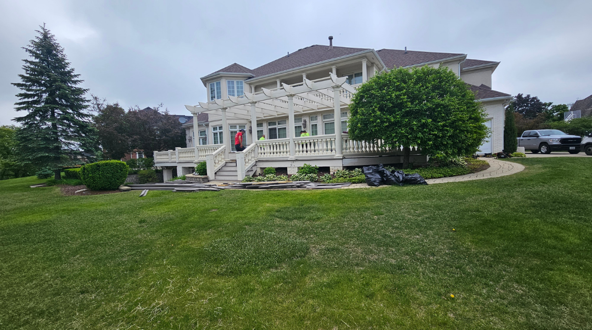 Large white historic house with wraparound porch on a green lawn, framed by trees and parked cars.