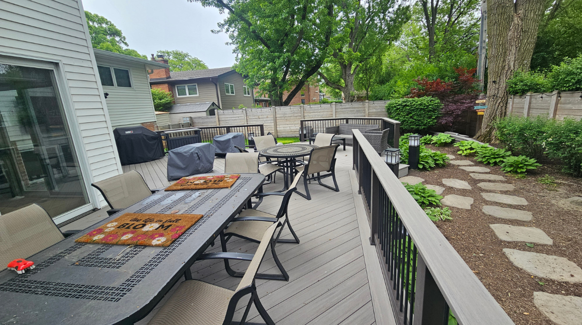 Outdoor deck with gray patio furniture, grill, and potted plants beside a landscaped yard