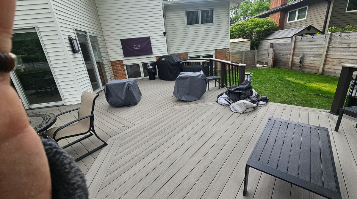 Backyard deck with gray furniture, grill, and storage bins beside a house and fenced lawn