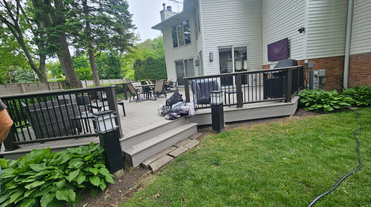 Backyard deck with black railings, steps, and a person sitting near a white house, surrounded by green lawn.