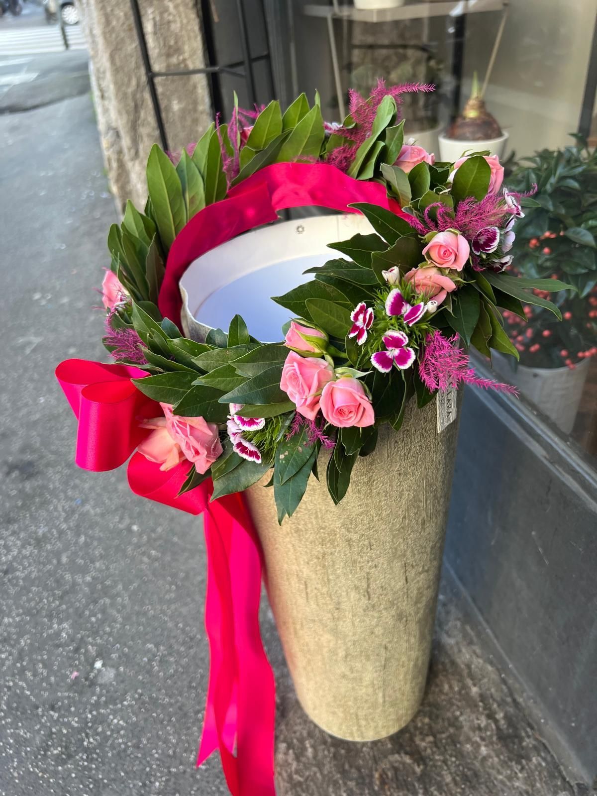 Composizione floreale rosa e bianca in un alto vaso dorato con un nastro rosa acceso accanto alla vetrina di un negozio