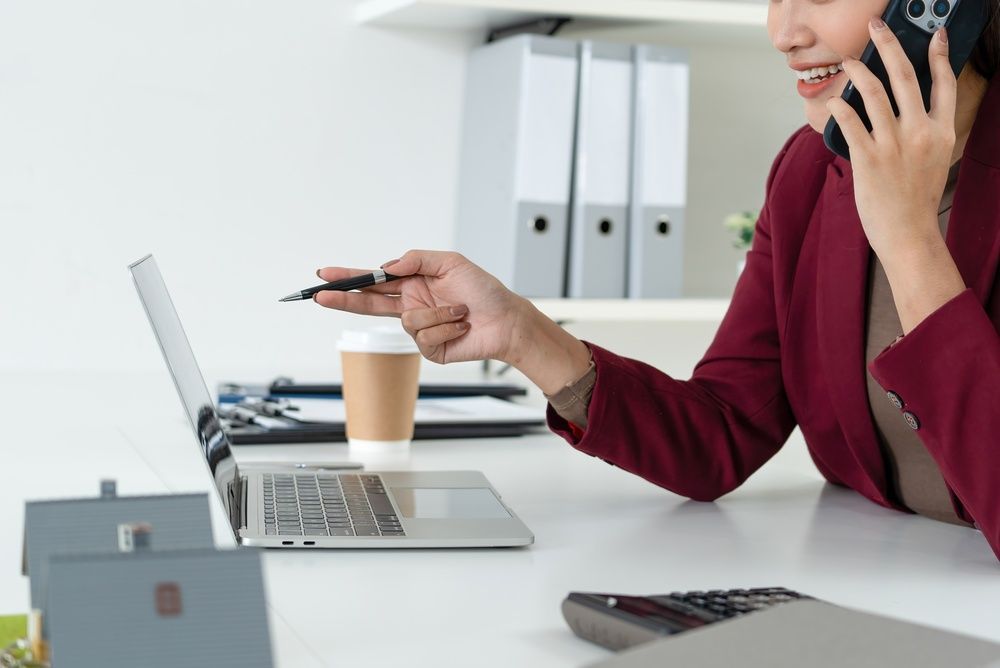Office worker on a phone call, pointing at a laptop on a desk with coffee and files.