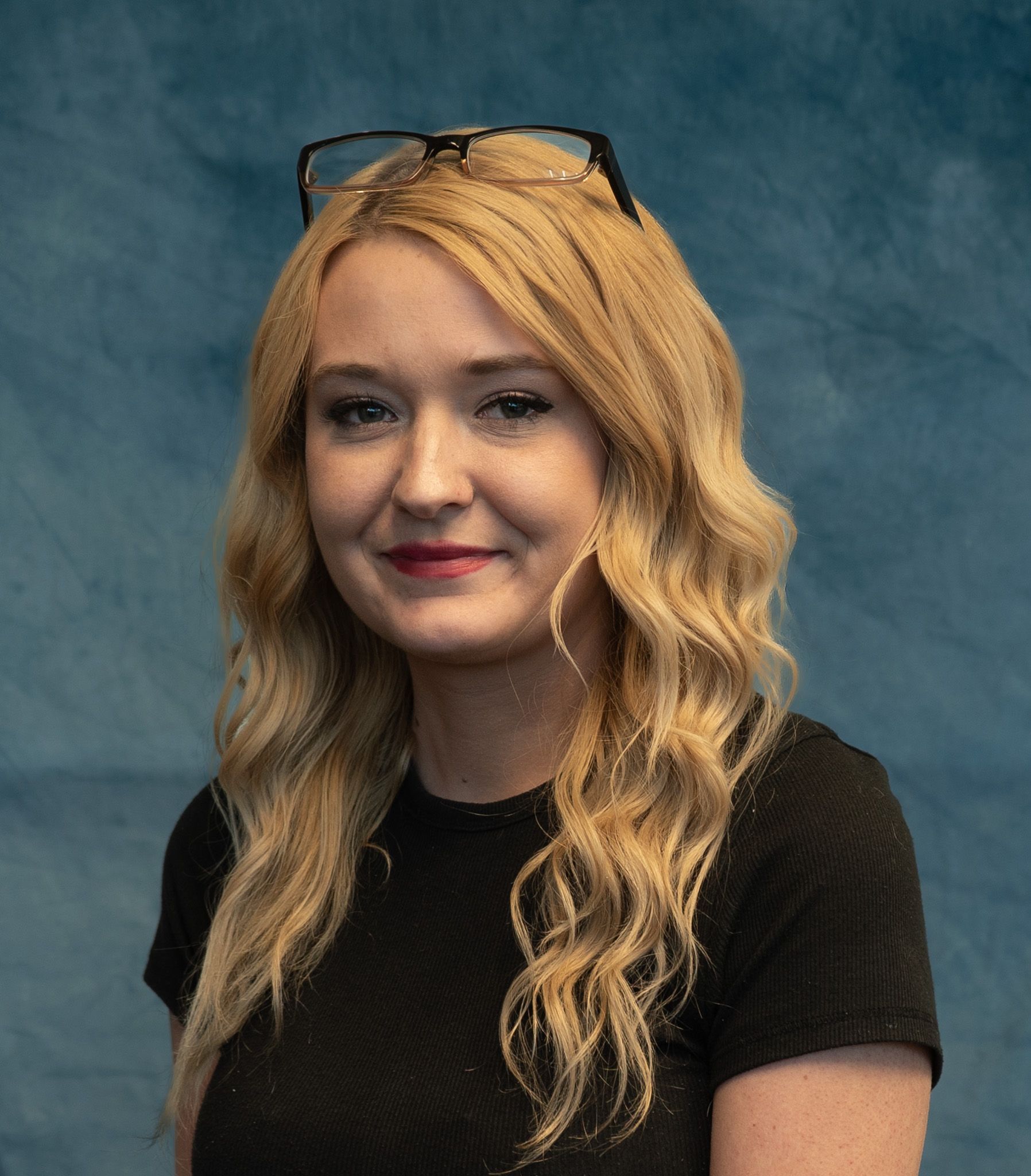 Blonde woman wearing glasses on her head, black top, smiling in front of a blue background.