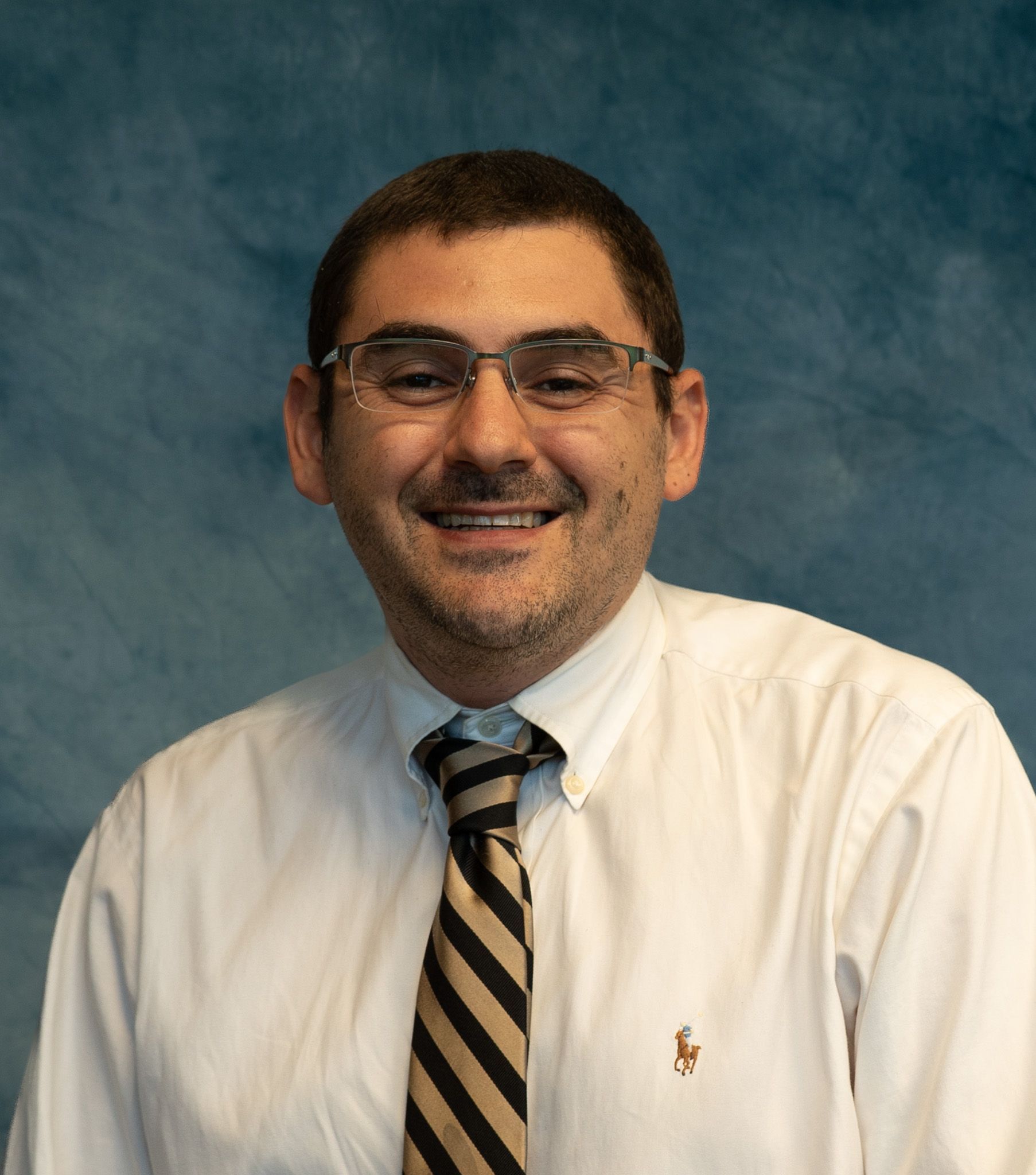 Man in a white shirt and tie with glasses smiling against a blue backdrop.