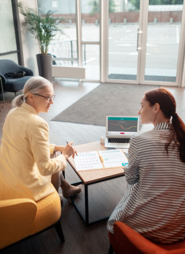 Two people in a bright office discuss documents across a table with a laptop.