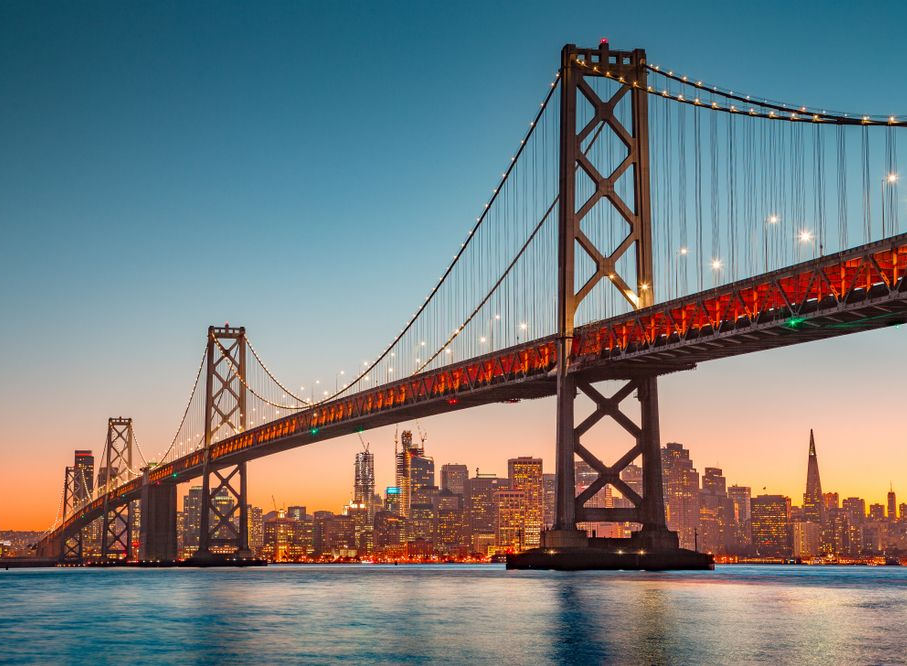 San Francisco Bay Bridge at sunset, spanning across the water with city skyline in the background.