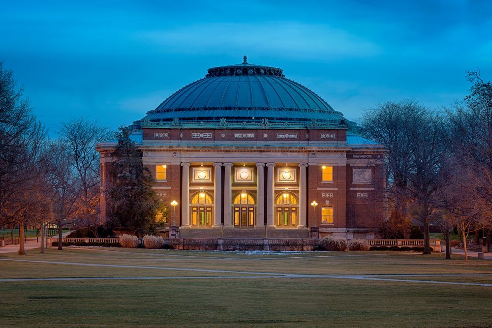 University of Illinois Library with lit windows at dusk, grass lawn, brick building, blue dome.