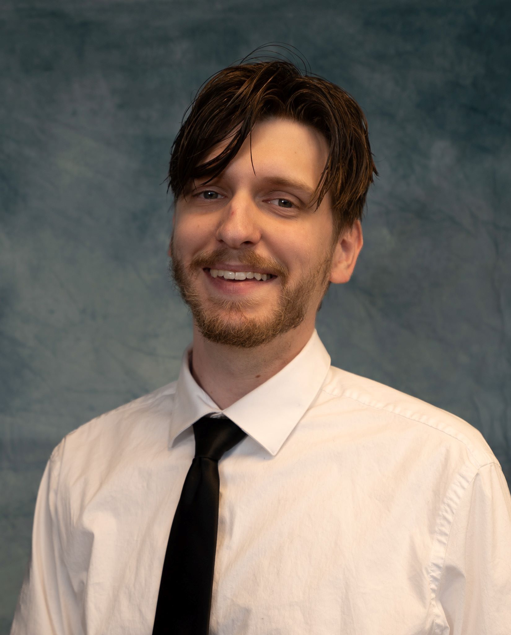 Man with brown hair and beard, wearing a white shirt and black tie, smiling in front of a blue background.
