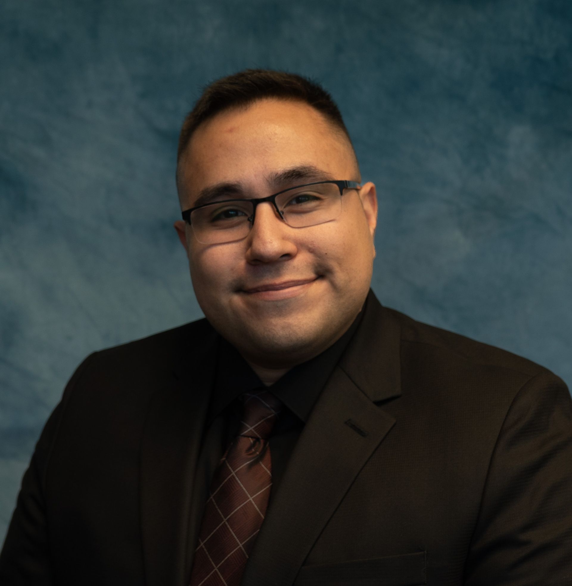 Man in a suit smiling, wearing glasses, against a blue backdrop.