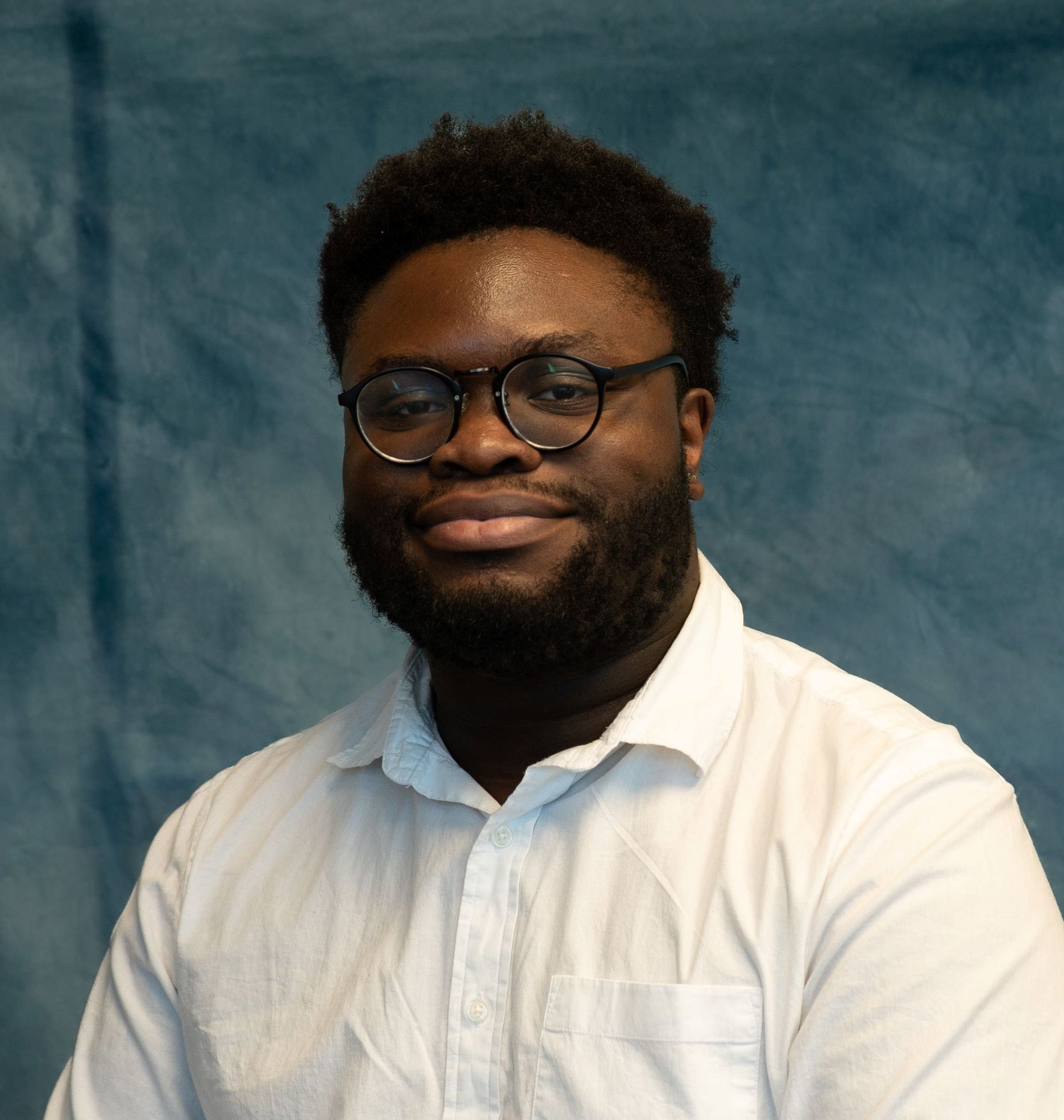 A Black man with a beard and glasses smiles; he wears a white shirt, stands in front of a blue backdrop.